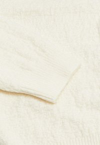 Dettaglio di un maglione bianco peloso con polsini a costine, caratterizzato da un motivo testurizzato ovunque, appariscente morbido e fibre che catturano la luce.