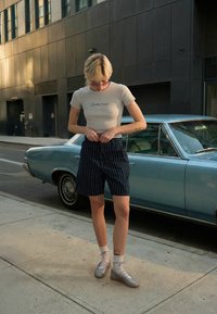 Poikaystävän tyyliset laivastonsiniset shortsit, joissa valkoiset pystysuorat raidat, yhdistettynä vaaleanbeigeen istuvaan t-paitaan ja harmaisiin nauhallisiin kenkiin. Moderni urbaani ympäristö.