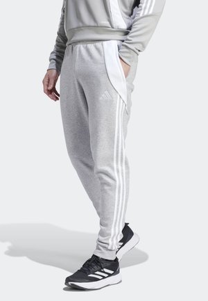 Person iført lysegrå Adidas træningsbukser med hvide striber, matchende top og sorte og hvide Adidas-sneakers, hånd i lommen.
