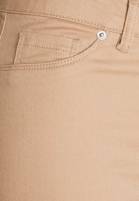 Beige denimbyxor har en liten framficka, silverfärgad metall, och en mjuk yta med synliga diagonala vävningsmönster.