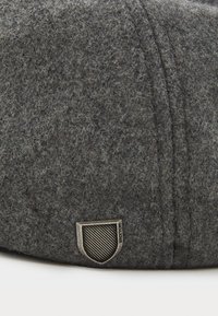 Gray felt cap with a textured finish, featuring a metal logo badge with a shield design on the side. Distinct seams run along the top.