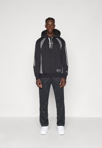 Mustapaitainen huppari harmailla yksityiskohdilla, jossa on naru, logo rinnassa ja yhteensopivat tummanharmaat collegehousut. Valkoiset lenkkitossut viimeistelevät asun.