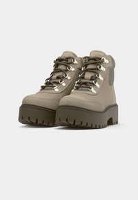 Beige plattform tursko med tykke brune såler, metallhull, mørkebrune lisser og polstrede ankelkrager.