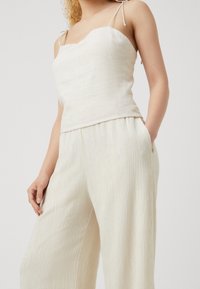 Top ligero de lino en blanco roto con tirantes finos y escote ondulado, combinado con pantalones de pierna ancha texturizados en color crema.