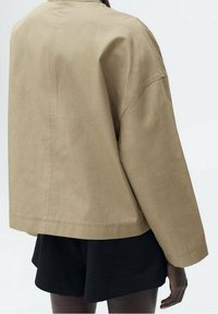 Khaki oversized jas met verlaagde schouders, rechte snit en minimale stiksels. Draag het over zwarte shorts met een matte textuur.