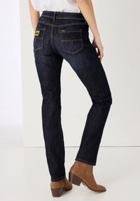 Jeans de mezclilla azul oscuro con un ajuste slim, que cuentan con bolsillos traseros y una etiqueta de marca amarilla en el bolsillo derecho. Se llevan con botas de tobillo marrones.