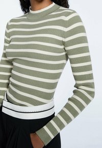 Groen en wit gestreepte ribgebreide top met een hoge hals en lange mouwen, voorzien van een witte riem om de taille met zwarte stiksels.