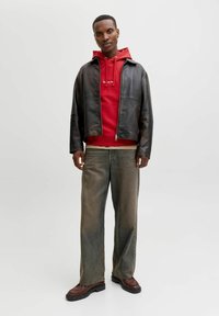 Veste en cuir noir, sweat à capuche rouge, t-shirt de couleur claire, jeans délavés à coupe décontractée et chaussures marron à semelles épaisses.