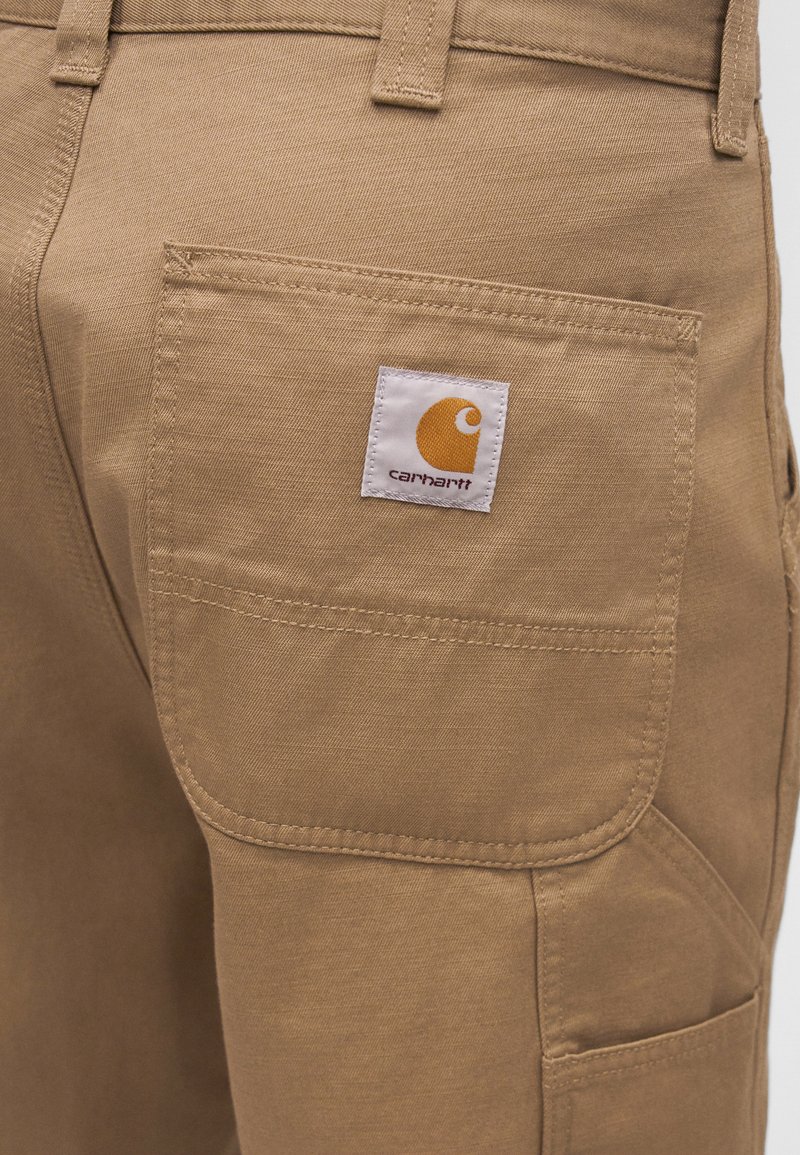 Gros plan sur la poche d'un pantalon Carhartt beige avec une étiquette de marque blanche et jaune visible cousue sur la poche.