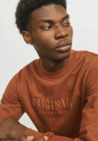 Sudadera de algodón marrón con el texto en relieve "ORIGINALS JACK & JONES". Cuello redondo, mangas largas. Textura suave con un diseño ajustado.