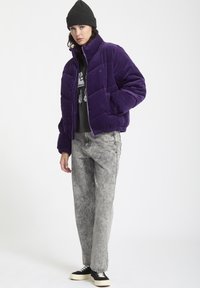 Paarse corduroy gewatteerde jas met een hoge kraag, ritssluiting en elastische manchetten. Draagt over een zwarte graphic sweatshirt en grijze jeans.