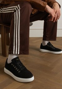 Sneakers in suede nera con suole bianche, caratterizzate da occhielli in metallo e dettagli con lacci. Indossati con pantaloni marroni con strisce bianche. Sfondo in legno.