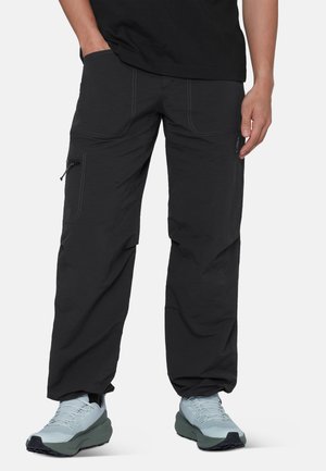 Mann trägt schwarze Cargo-Hose mit Reißverschlusstasche und hellgraue Sportschuhe, steht vor einem schlichten Hintergrund.
