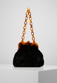 Černá plyšová kabelka s řetízkem a uzávěrem z akrylového želvovitého vzoru. Hladký kulatý tvar s měkkou texturou, umístěná na bílém podstavci.