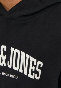 Sudadera negra con el texto de la marca en blanco "JONES" y "since 1990". Cuenta con capucha con cordón y una tela suave y texturizada. Enfoque en el detalle del cuello.