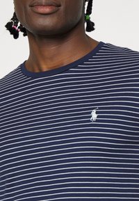 Modré a bílé pruhované tričko s kulatým výstřihem a malým vyšitým logem na levé hrudi. Bavlněný materiál, hladká textura.