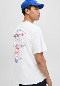 Fehér pamut póló, amelyen retro grafikai nyomat található "Abarc Hotel" felirattal piros és kék színekben, díszítő elemekkel és szöveggel a hátsó oldalon.