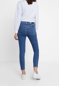 Jeans ajustados en denim azul medio, de tiro alto, con una textura suave. Presenta dos bolsillos traseros y detalles de costura mínimos. Combinados con una sudadera blanca estampada y zapatillas blancas.