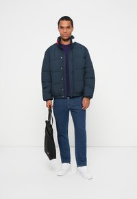 Chaqueta acolchada azul marino con botones a presión, que presenta un cuello rojo y blanco. Combinada con jeans oscuros, zapatillas blancas y una bolsa de mano negra.