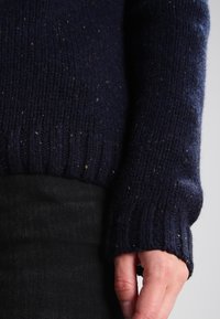 Marineblauer Strickpullover mit strukturiertem Finish, ausgestattet mit gerippten Bündchen und mehrfarbigen Sprenkeln. Über schwarzer Denim getragen.
