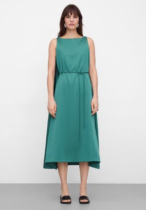 Vrouw in mouwloze teal midi-jurk met dunne riem, recht afgewerkte zoom aan de voorkant, en open zwarte slippers, staand tegen een witte achtergrond.