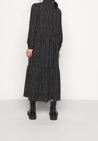 Černé dlouhé šaty s volánkovou sukní, zdobené bílými vertikálními pruhy, s pevně tvarovaným límcem a nošené s černými chunky botami.