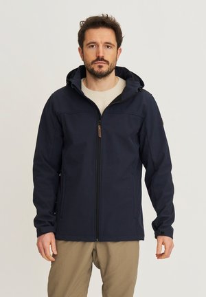 Man draagt een marineblauwe ritsvest met capuchon over een beige trui en kaki broek, staand tegen een effen lichte achtergrond.