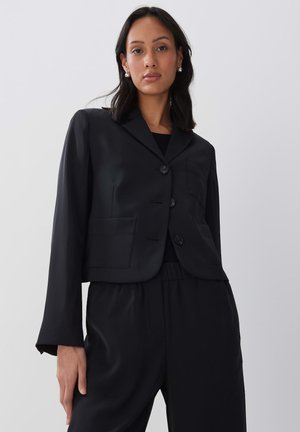 Black cropped blazer made of smooth fabric, featuring a notched collar, three buttons, and two front pockets. Styled with matching black pants.