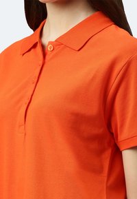 Frau trägt ein leuchtend orangefarbenes Polohemd mit kurzen Ärmeln, einem umgeschlagenen Kragen und vier Knöpfen, gezeigt von Hals bis zur Mitte des Oberkörpers.