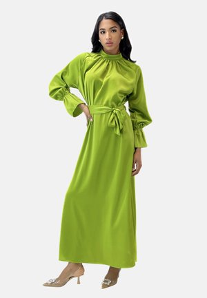 Robe en satin vert longue avec un col haut, des manches bouffantes longues et une taille à nouer. Portée avec des chaussures à talons transparents ornées de détails.