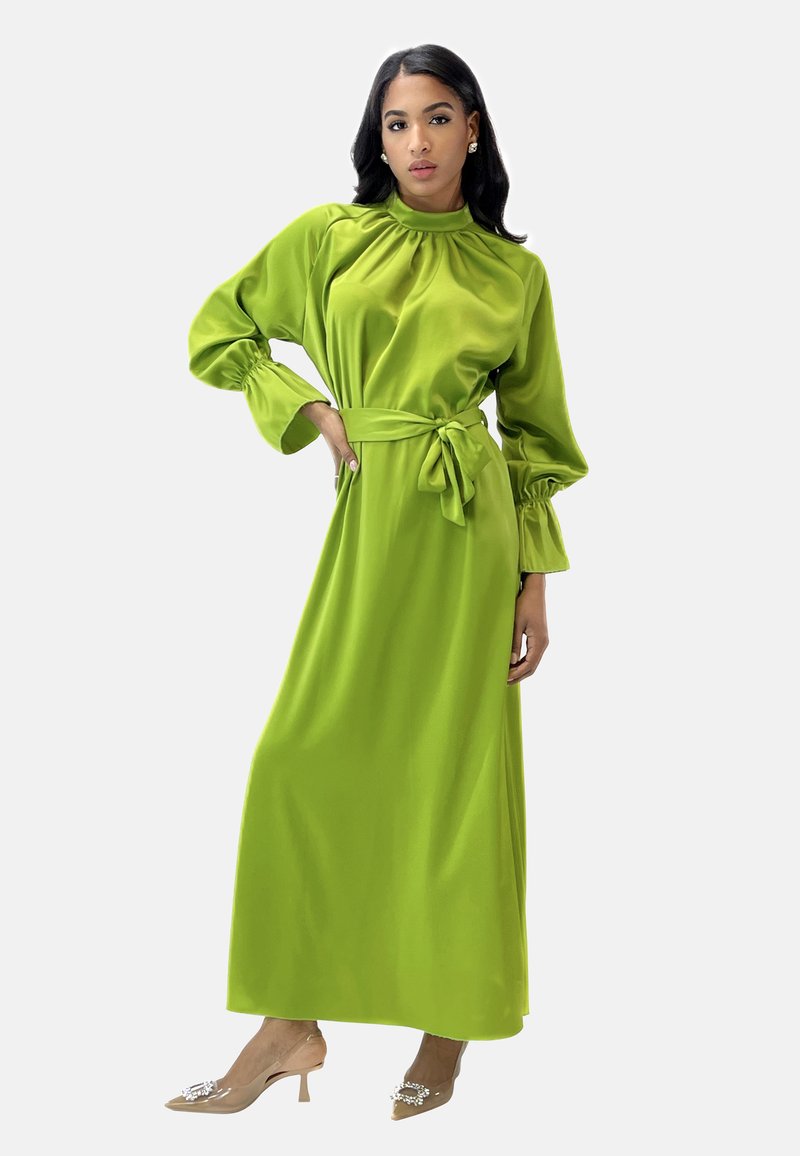 Langes, grünes Satin-Kleid mit hohem Kragen, langen Puffärmeln und einer Taillenschnürung. Getragen mit transparenten Keilabsätzen, die mit Verzierungen ausgestattet sind.