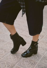 Czarne lakierowane botki za kostkę z grubym obcasem, zamkiem błyskawicznym i akcentem w postaci klamry. Nosi się je z przyciętymi, ciemnoniebieskimi dżinsami.