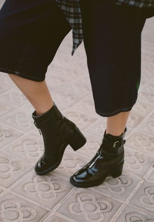 Stivaletti in pelle verniciata nera con tacco spesso, chiusura a zip e un dettaglio con fibbia. Indossati con pantaloni jeans scuri cropped.