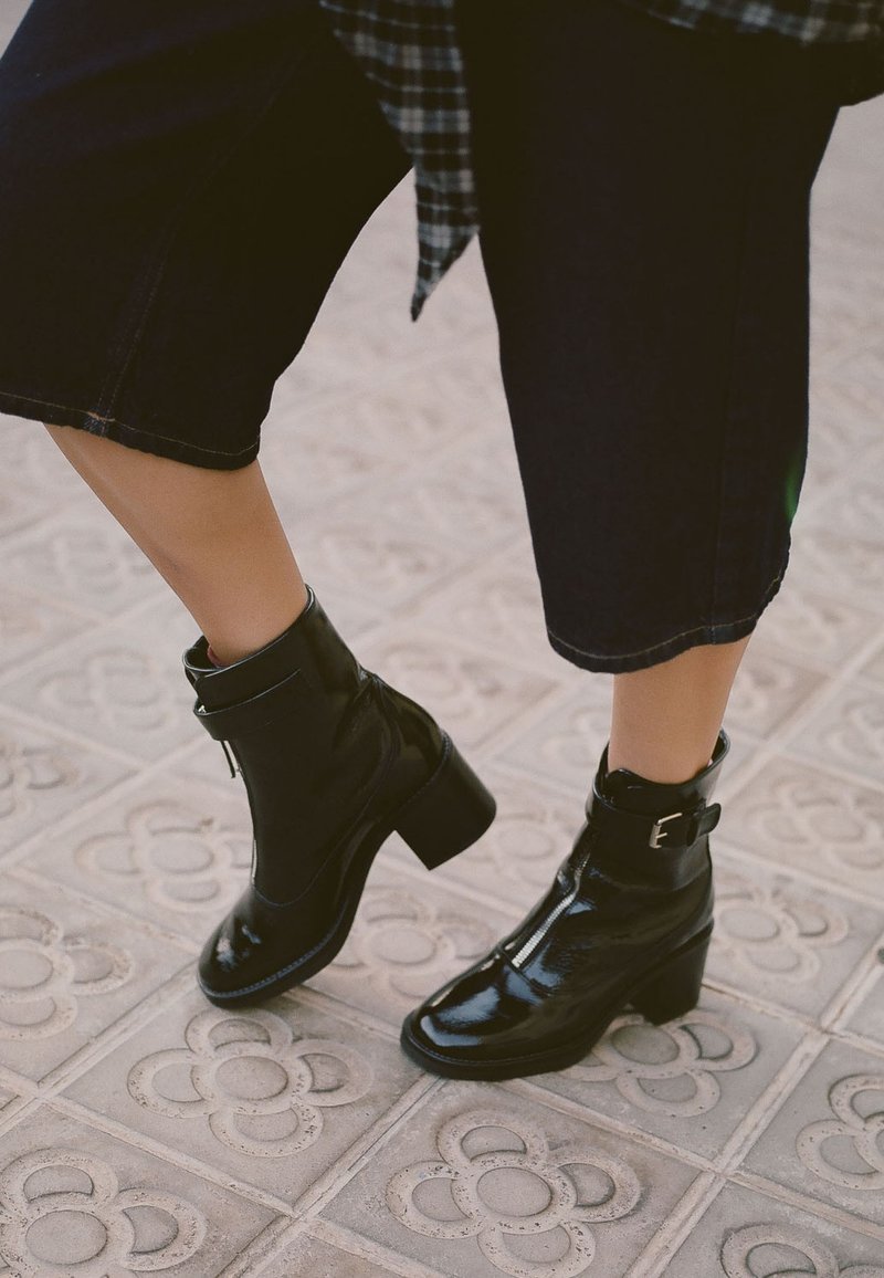 Czarne lakierowane botki za kostkę z grubym obcasem, zamkiem błyskawicznym i akcentem w postaci klamry. Nosi się je z przyciętymi, ciemnoniebieskimi dżinsami.