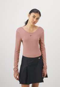 Hosszú ujjú, bordázott rózsaszín felső széles dekoltárral és fekete Nike logóval, fekete, bővülő szoknyával párosítva, amely színes folttal rendelkezik.