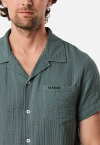 Camicia verde a maniche corte con texture, colletto a risvolto, bottoni bianchi e taschino sul petto ricamato con la scritta "SEASIDE".