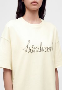 Femme portant un t-shirt ample jaune pâle à manches courtes avec "handvcref" écrit en cursive sur la poitrine.