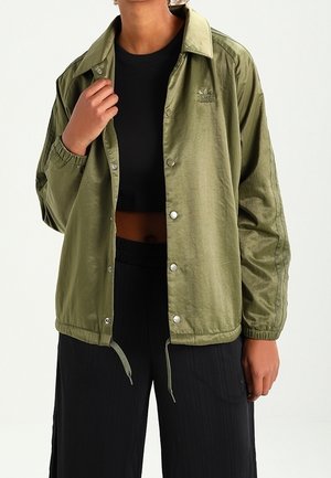 Person trägt eine olivgrüne, knöpfbare Jacke über einem schwarzen Crop-Top und schwarzen Hosen, eine Hand hält den Jackenkragen.