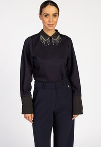 Marineblaues Oberteil mit schwarzem Kragen und verzierten Perlendetails, kombiniert mit marineblauen, hoch taillierten Hosen mit Knopfdetails an den Ärmeln.