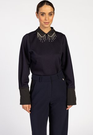 Marineblauw top met zwarte kraag en versierde kraaldetails, gecombineerd met marineblauwe high-waisted broek met knopenaccenten op de mouwen.