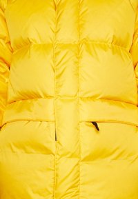 Veste matelassée jaune avec un design piqué, comportant des motifs horizontaux et verticaux, une poche zippée, et une texture légère et lisse.