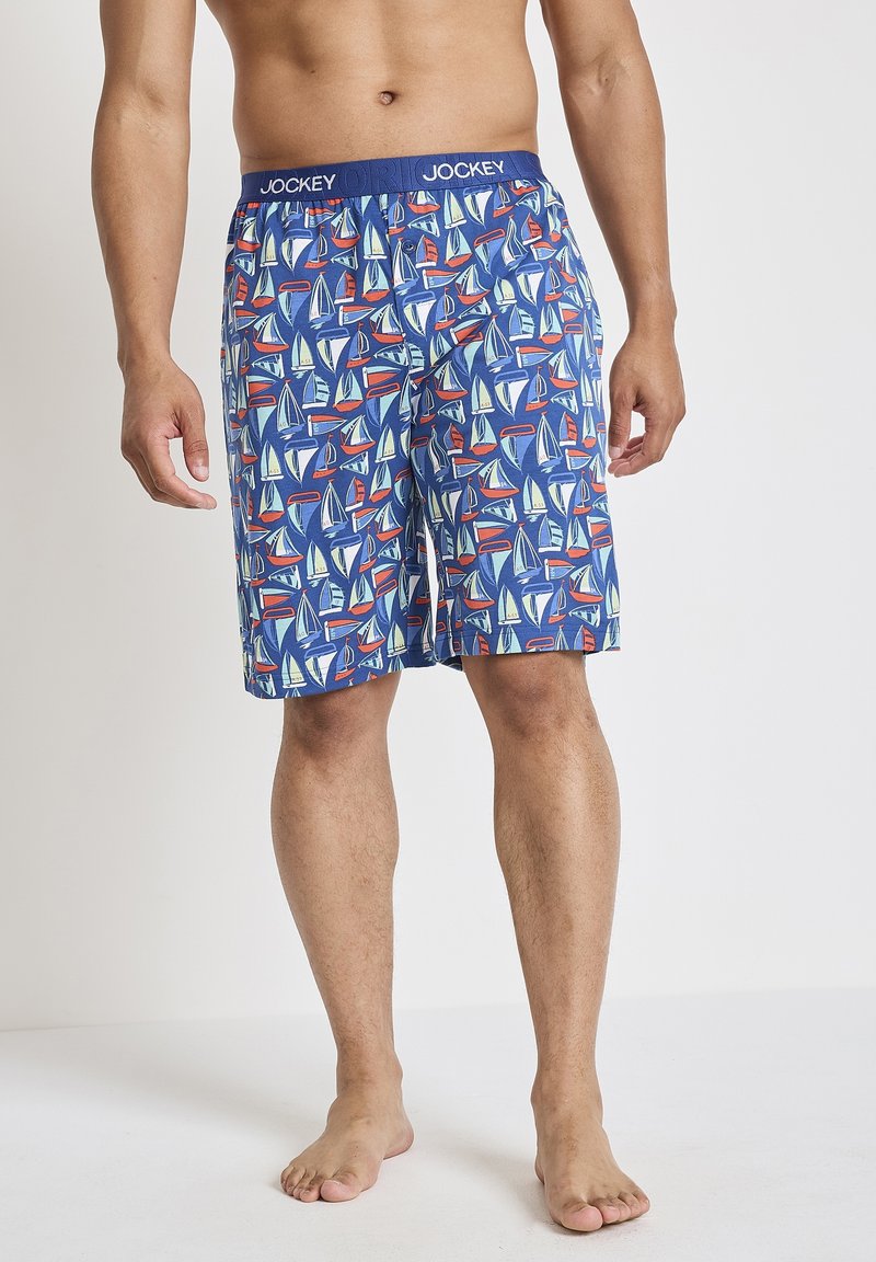 Uomo che indossa pantaloni shorts blu con motivi di barche a vela colorate e una cintura elastica con marchio, in piedi a piedi nudi su un pavimento bianco.