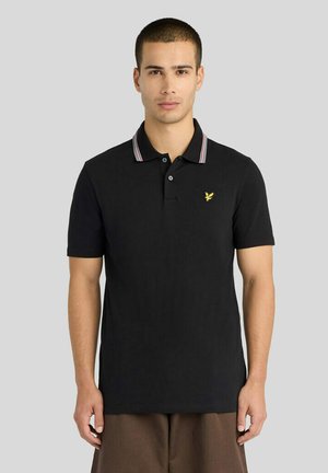 Ung mand iført en sort kortærmet polo med et lille gult emblem og stribet krave står foran en ensfarvet grå baggrund.