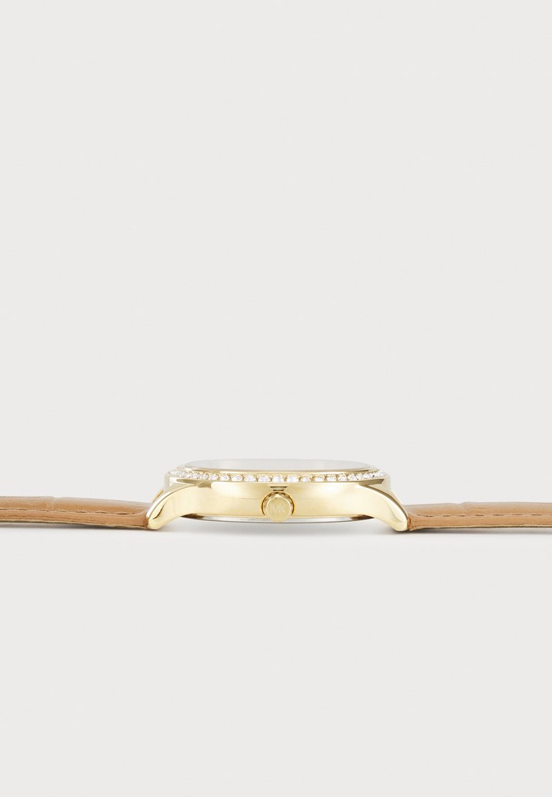 Montre dorée avec un boîtier rond, accents en cristal sur la lunette, et un bracelet en cuir marron clair. Pas de marques visibles sur le bracelet.