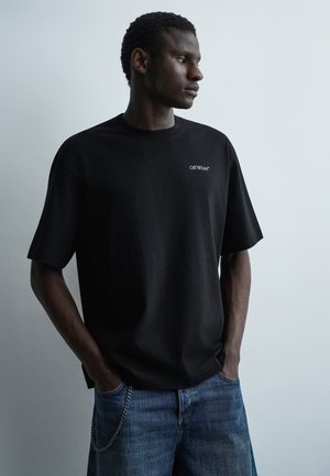 Άνδρας με χέρια στις τσέπες, φοράει μαύρο T-shirt Off-White και μπλε τζιν, κοιτάζοντας δεξιά σε απλό φωτεινό φόντο.