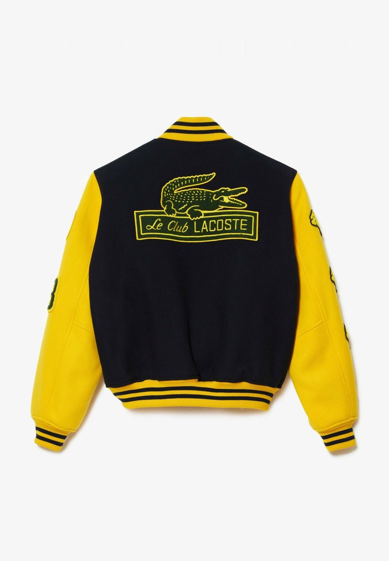 Veste de varsity noire et jaune avec des poignets côtelés, présentant un logo de crocodile brodé et le texte "Le Club Lacoste" au dos.