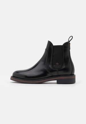 Bota Chelsea de cuero negro liso con panel lateral elástico, ribete de suela marrón, tacón bajo y lengüeta con marca en la parte trasera.