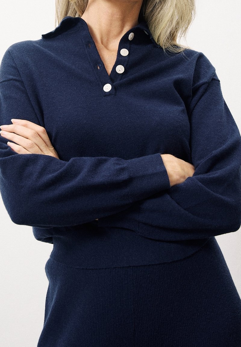 Personne les bras croisés portant un pull bleu marine à manches longues avec un col en V boutonné.
