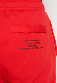 Röda Nike sweatpants har en bakficka med en svart tryckt logotyp och textdetaljer. Materialet verkar mjukt med en slät textur.