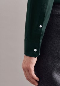 Lewe ramię w ciemnozielonej, długiej koszuli z białymi guzikami, opoczywające obok szarych wełnianych spodni.